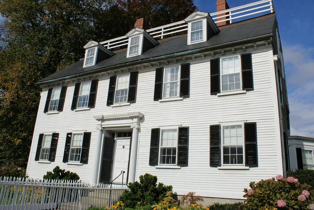 A white clapboard colonial house with black shutters and dormers on a dark shingle roof