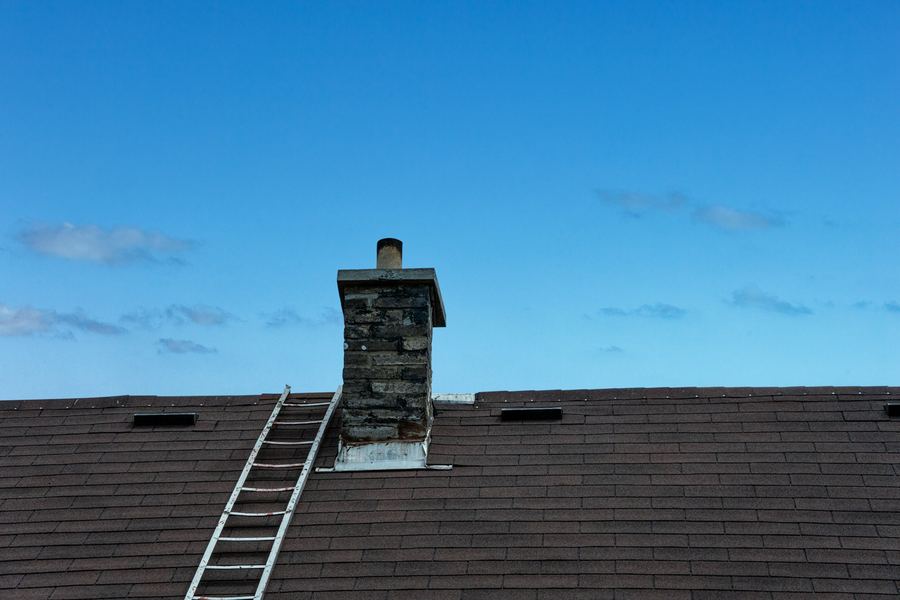 An aluminum ladder resting on a dark asphalt shingle roof beside a stone chimney, under a blue sky